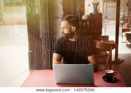 Young Bearded Businessman Wearing Black Tshirt Working Laptop Urban Cafe.Man Sitting Wood Table Cup Coffee Looking Through Window.Coworking Process Business Startup.Blurred Background.Sunlight effect