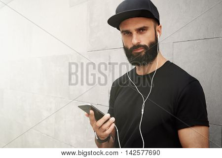 Bearded Muscular Man Wearing Black Tshirt Blank Snapback Cap Summer Time.Young Men Smiling Opposite Empty Gray Concrete Wall Background Using Smartphone Headphones.Horizontal Mockup.Color Filter