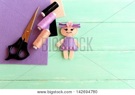 Handmade cute doll, scissors, thread, needle, felt sheets on wooden background with blank space for text. How simply and cheaply make a children toy. Creative doll crafts with felt for kids