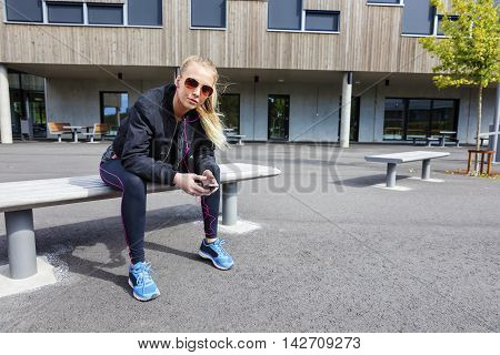 Beautiful woman with sunglasses sits on a bench in the city wearing a jacket and workout outfit. Listens to music and using smart phone.