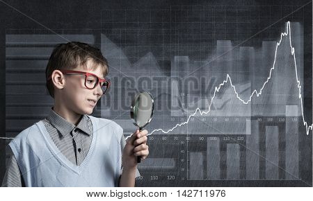 Cute boy looking through magnifying glass against graphs at chalkboard