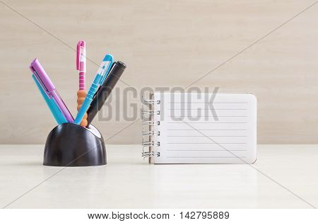 Closeup color pen with black ceramic desk tidy for pen and white page with line at the note book on blurred wood desk and wood wall textured background
