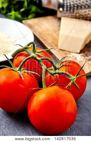 Branch of fresh red tomatoes with water drops on and parmesan or parmigiano reggiano cheese on back with grater and grated cheese on white plate and mint beam. Concept of italian cuisine ingredients