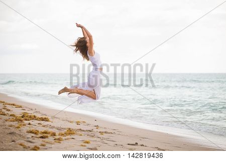 Young woman in a white dress jumping on the beach by the ocean. Happy vacation. Beach girl has fun by ocean. Excited girl jumping on white sand by beautiful blue sea.