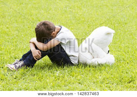 Sad disappointed little boy sitting with a teddy bear. Both turned away. Both lowered their heads. Outdoor. Sadness fear frustration loneliness concept