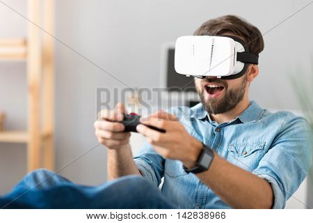 Pleasant time spending . Cheerful handsome man holding game console and playing video games while wearing virtual reality device
