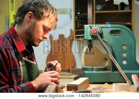 A young man in a flannel shirt is working as a luthier using tools to building a guitar in his home workshop.