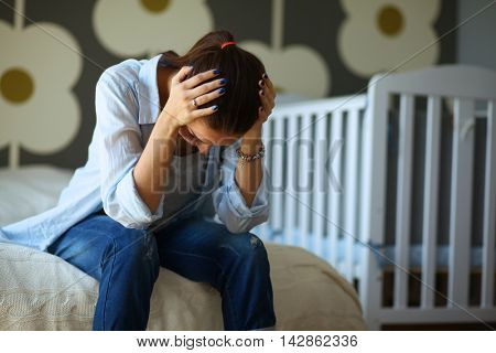Young tired woman sitting on the bed near children's cot.