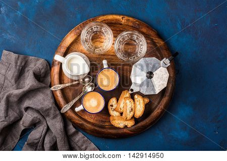 Coffee espresso in cups with italian cantucci, cookies and milk in jug on wooden serving round board over dark blue painted plywood background, top view