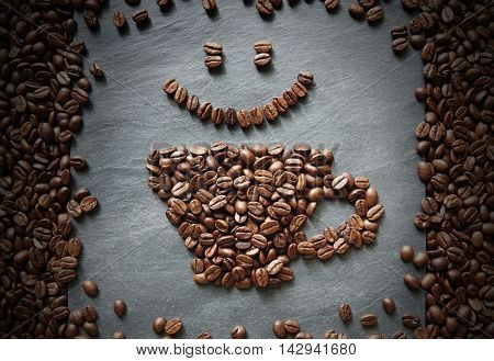 Cup of coffee and smiley face from coffee beans. Heap of coffee bean in form of cup on grey stone surface texture. Coffee shop or cafe background. Soft color toning, top view
