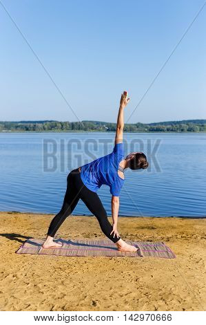Yoga girl in triangle pose on the river bank. Young woman practicing yoga on mat outdoors. Healthy lifestyle. Yoga. Triangle or trikonasana pose of yoga asana. Young fit woman doing yoga triangle pose