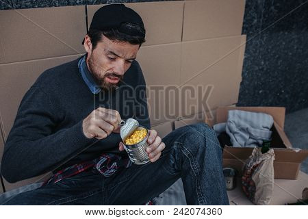 Poor And Hmeless Man Is Sitting On Cardboard And Opening Can Of Corn. He Is Looking At It. Guy Is St