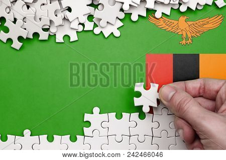 Zambia Flag  Is Depicted On A Table On Which The Human Hand Folds A Puzzle Of White Color.