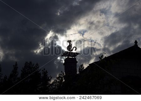 A Stuffed Cat Dummy Sits On A Chimney At Home, Thick Black Clouds And Sun, Chimney And Cat
