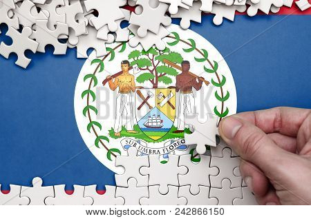 Belize Flag  Is Depicted On A Table On Which The Human Hand Folds A Puzzle Of White Color.