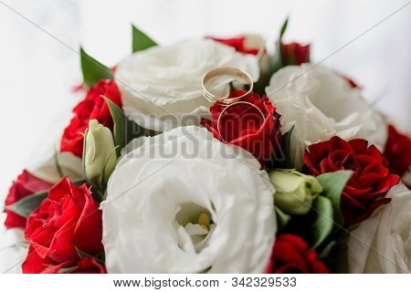Gold Wedding Rings Of The Bride And Groom On Red Roses And White Eustoms Close Up. Symbol Of Love An