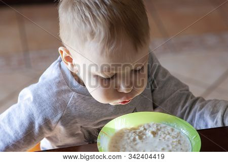 Cute Caucasian Baby Boy With Blond Hairs Blows Into The Porridge At The Table, In Sunny Morning. Chi