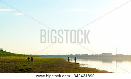 A Group Of Asian Yakut Men And Women With Bags And Buckets Walk Along The Stone Wild Bank Of The Nor