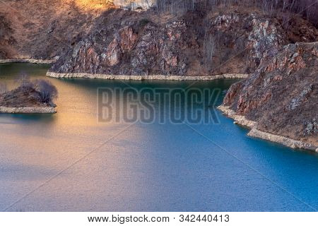 The First Rays Of The Sun Illuminate A Lake With Rocky Shores And Create Glare On A Calm Surface Of 