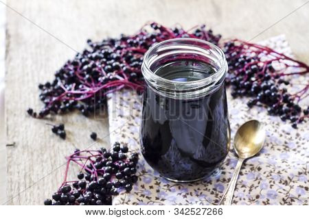 Homemade Black Elderberry Syrup In Glass Jar And Bunches Of Black Elderberry In Background