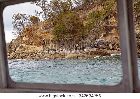 Rocky Coastal Shoreline With Brown Layered Volcanic Shale And Spindly Trees And Sparse Vegetation, A
