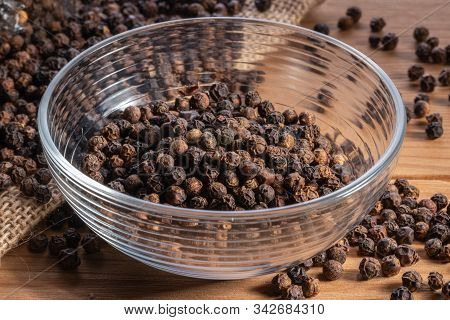 Black Pepper Grains In A Transparent Deep Dish On A Wooden Surface. In The Background, Sackcloth And