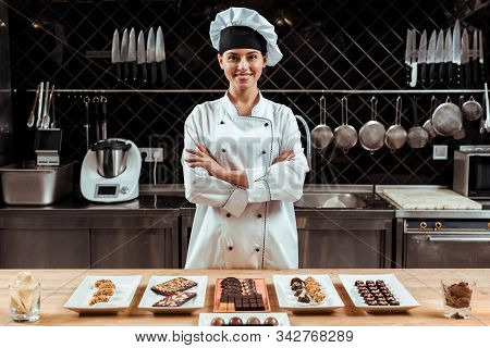 Happy Chocolatier In Chef Hat Standing With Crossed Arms Near Tasty Chocolate Candies On Plates