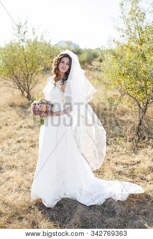 Beautiful Smiling Bride Wearing A Natural Flower Wreath And Standing In The Golden Summer Fields Wit