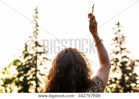 A Caucasian Woman Is Viewed From Behind As She Raises A Piece Of Tree Bark To The Sky During Sundown
