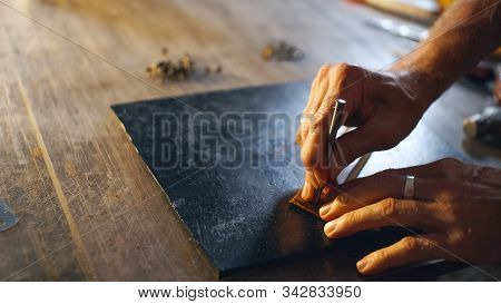 Leatherwork Concept. Tanner Makes Hole In Workpiece. Male Hands Close Up Holding Tool.