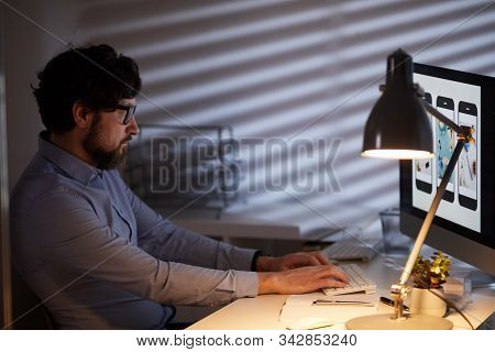 Serious Young Man Sitting At The Table In Front Of Computer Monitor And Typing On Computer Keyboard 