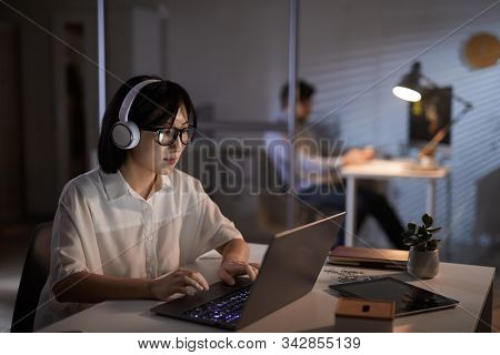 Serious Asian Designer In Headphones Working On Laptop Computer At The Office Till Late Evening