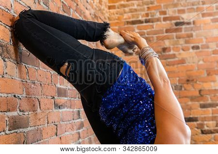 A Closeup View Of An Upside Down Woman, Using A Red Brick Wall To Rest Her Knee Against As She Stret