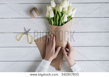 Woman Florist Wrapping Beautiful Bouquet Of White Tulips In Pack Craft Paper On The Wooden Table. Fl
