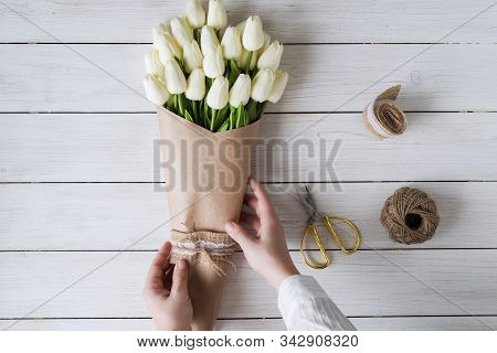Woman Florist Wrapping Beautiful Bouquet Of White Tulips In Pack Craft Paper On The Wooden Table. Fl