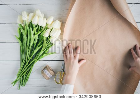 Woman Florist Wrapping Beautiful Bouquet Of White Tulips In Pack Craft Paper On The Wooden Table. Fl