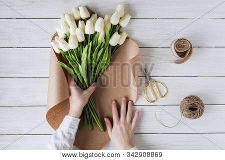 Woman Florist Wrapping Beautiful Bouquet Of White Tulips In Pack Craft Paper On The Wooden Table. Fl