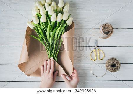 Woman Florist Wrapping Beautiful Bouquet Of White Tulips In Pack Craft Paper On The Wooden Table. Fl