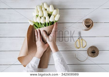 Woman Florist Wrapping Beautiful Bouquet Of White Tulips In Pack Craft Paper On The Wooden Table. Fl