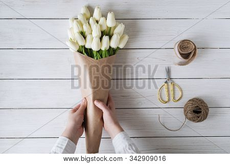Woman Florist Wrapping Beautiful Bouquet Of White Tulips In Pack Craft Paper On The Wooden Table. Fl