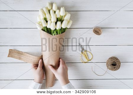 Woman Florist Wrapping Beautiful Bouquet Of White Tulips In Pack Craft Paper On The Wooden Table. Fl