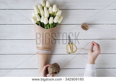 Woman Florist Wrapping Beautiful Bouquet Of White Tulips In Pack Craft Paper On The Wooden Table. Fl