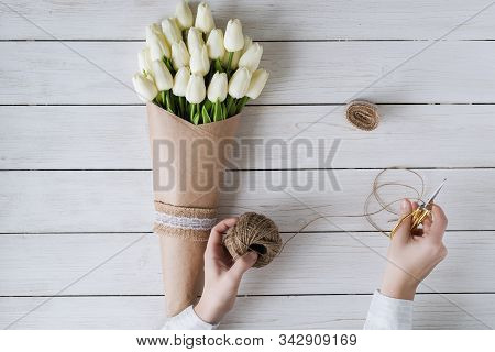 Woman Florist Wrapping Beautiful Bouquet Of White Tulips In Pack Craft Paper On The Wooden Table. Fl
