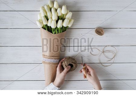Woman Florist Wrapping Beautiful Bouquet Of White Tulips In Pack Craft Paper On The Wooden Table. Fl
