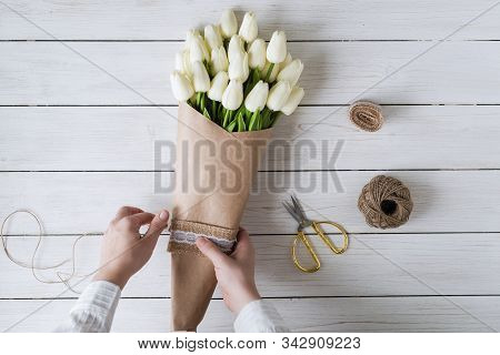 Woman Florist Wrapping Beautiful Bouquet Of White Tulips In Pack Craft Paper On The Wooden Table. Fl