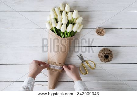 Woman Florist Wrapping Beautiful Bouquet Of White Tulips In Pack Craft Paper On The Wooden Table. Fl