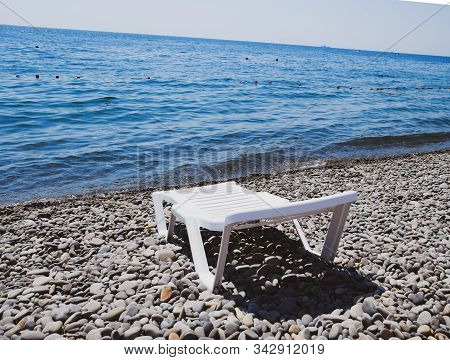Chaise Lounge Is White On A Stony Beach By The Coastline. Chaise Lounge By The Sea.