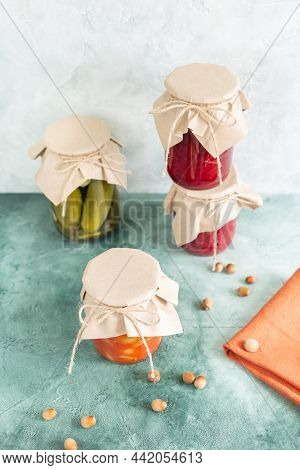 Pickles, Canned Vegetables In Glass Jars And Scattered Nuts And Terracotta Napkin On A Green Texture