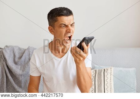Horizontal Shot Of Dark Haired Young Adult Male Wearing White Casual Style T Shirt Sitting On Sofa, 