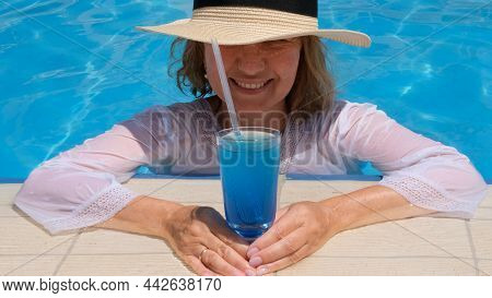 Smiling Senior Woman Relaxing Near The Blue Outdoor Swimming Pool With A Blue Cocktail. People Are E